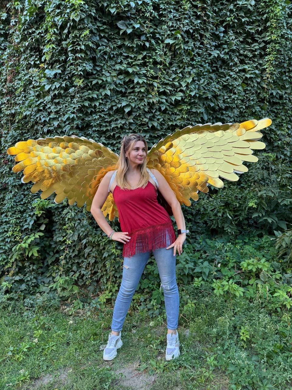 May include: A person wearing large, golden, layered wings, posing in front of a wall of green foliage. The wings have a textured, metallic appearance. The person is wearing a red top, blue jeans, and white sneakers.