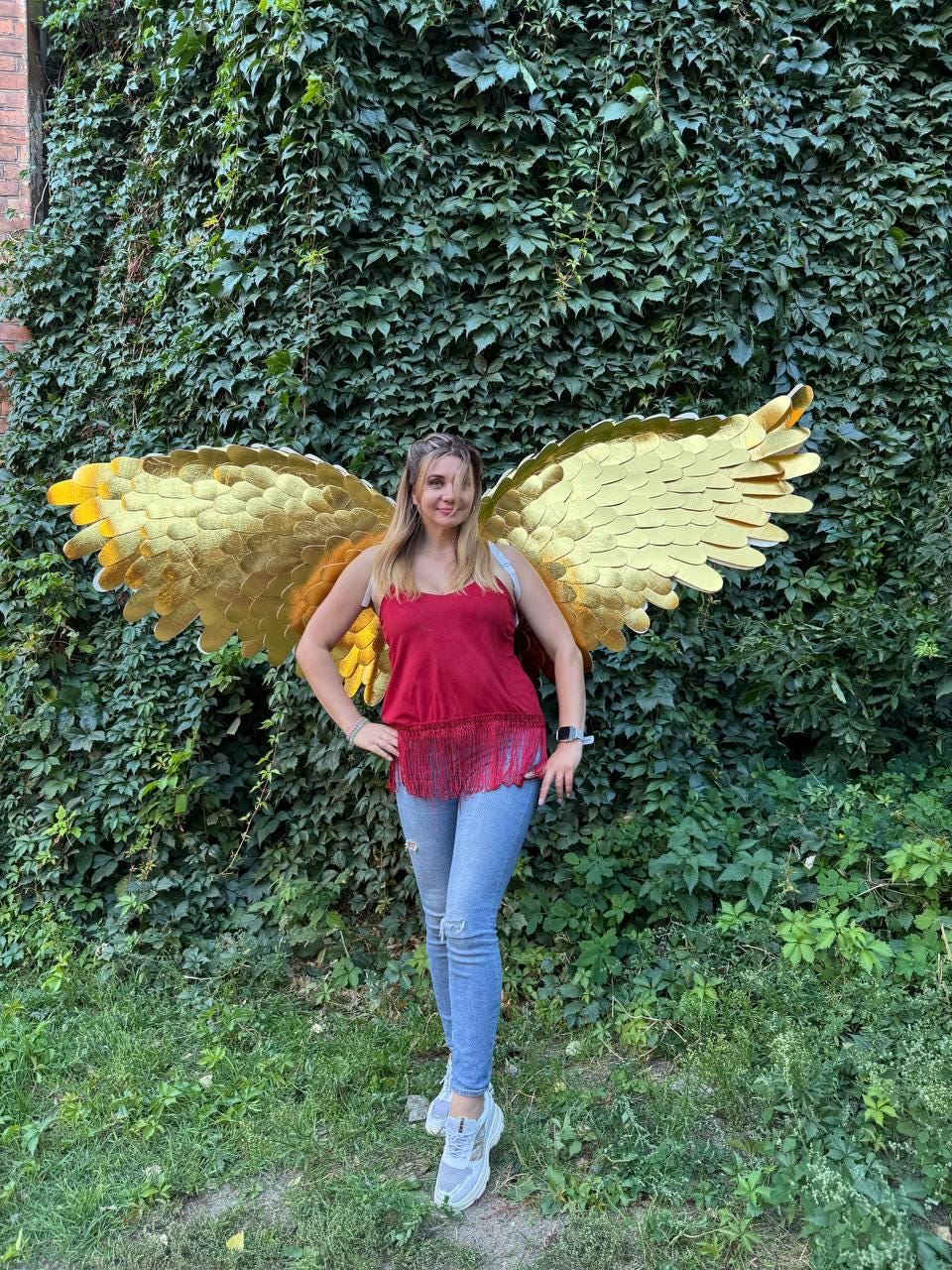 May include: A person wearing gold-colored angel wings, a red top, and blue jeans stands in front of a wall of green foliage. The wings are large and detailed, with a layered design. The person is smiling.
