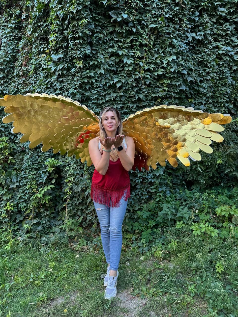 May include: A woman wearing large, golden angel wings stands in front of a wall of green foliage. The wings are detailed with layered feathers and have a gradient of gold and red. She is wearing a red top, blue jeans, and gray sneakers.
