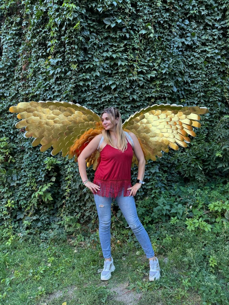 May include: A woman wearing gold-colored angel wings, a red top with fringe, and blue jeans stands in front of a wall of green foliage. The wings are large and detailed, with a layered design.