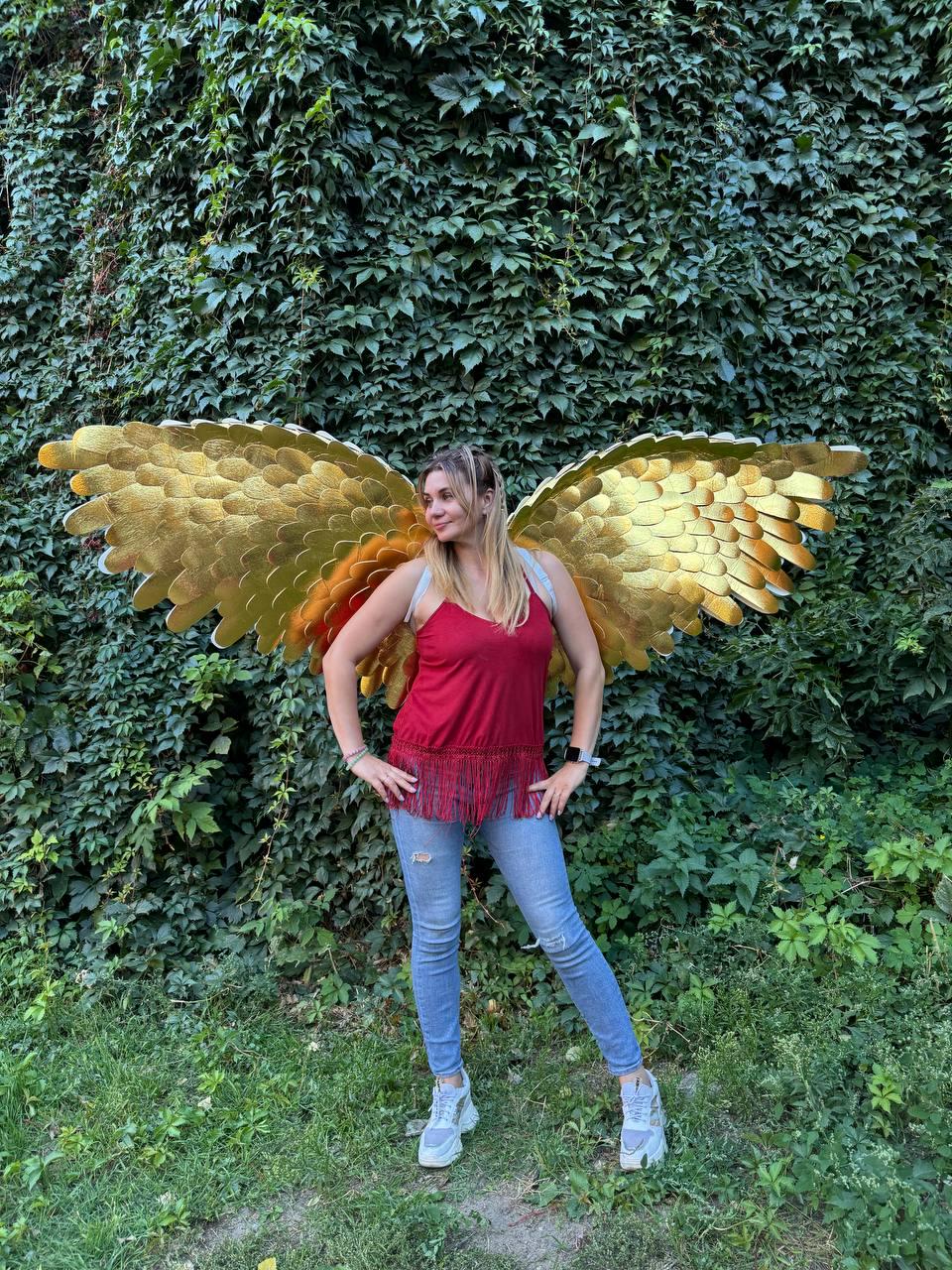 May include: A woman wearing gold-colored angel wings, a red top with fringe, and blue jeans stands in front of a wall of green foliage. The wings are large and detailed, with a layered design.