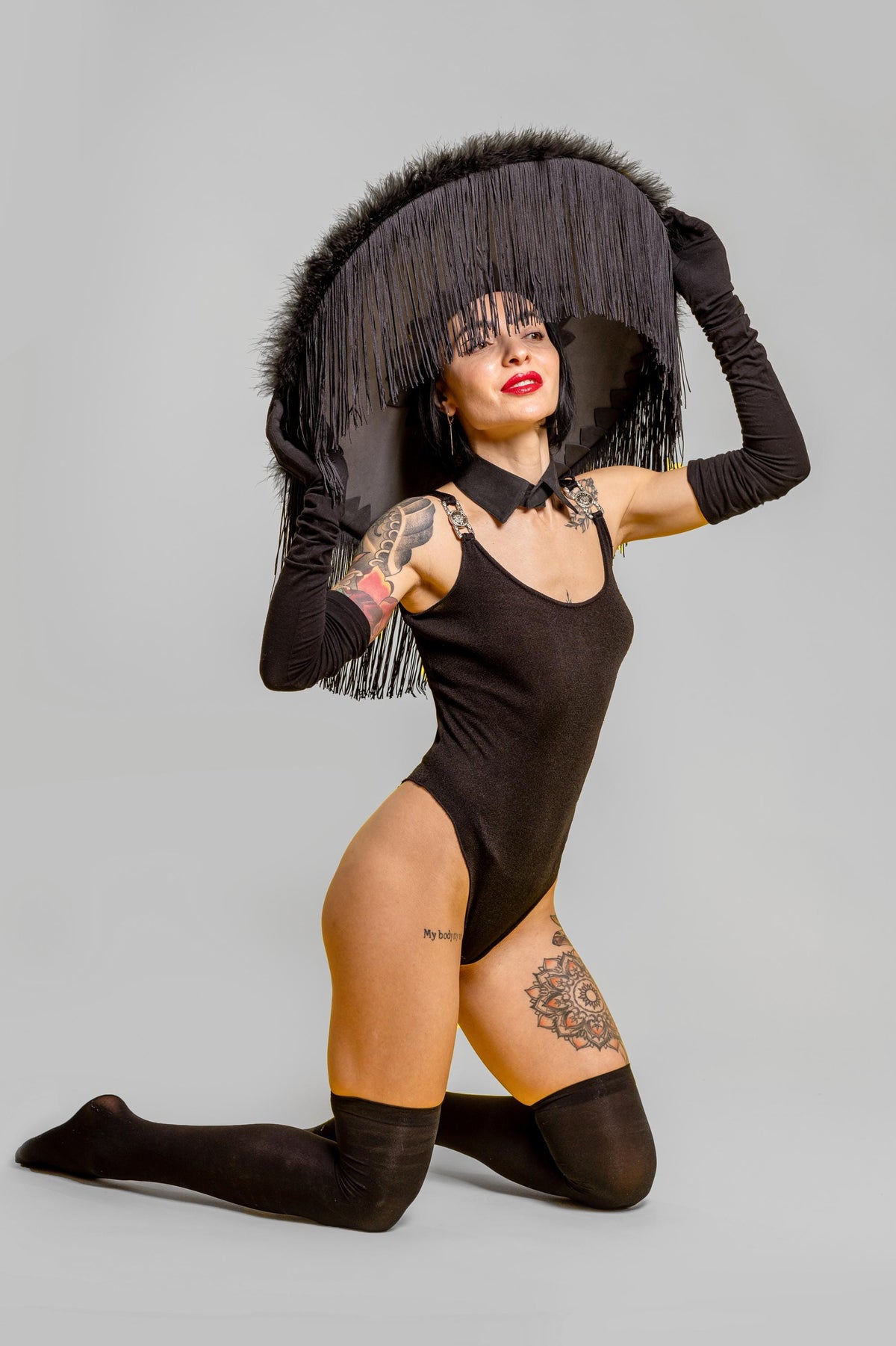 May include: A woman kneels, wearing a black bodysuit, thigh-high stockings, and long gloves. She holds a large, black hat with fringe. The hat and gloves create a dramatic, theatrical look. Tattoos are visible on her arm and leg.