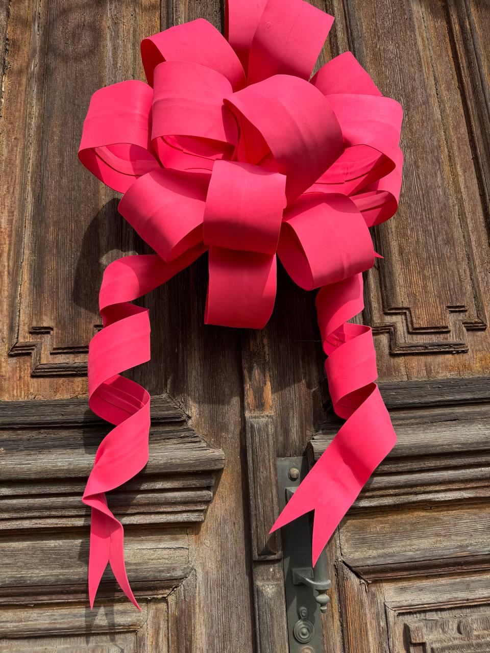 May include: A large, vibrant pink bow made of paper or fabric, attached to a wooden door. The bow has multiple loops and long, curling streamers. The door is made of dark wood with decorative carvings and a metal handle.
