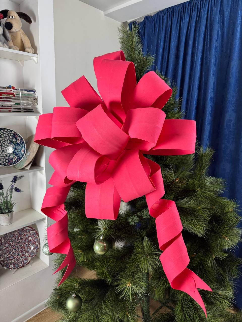 May include: A large, vibrant pink bow adorns the top of a green Christmas tree. The bow's ribbon cascades down in elegant spirals. The tree is decorated with small green ornaments, creating a festive holiday display.