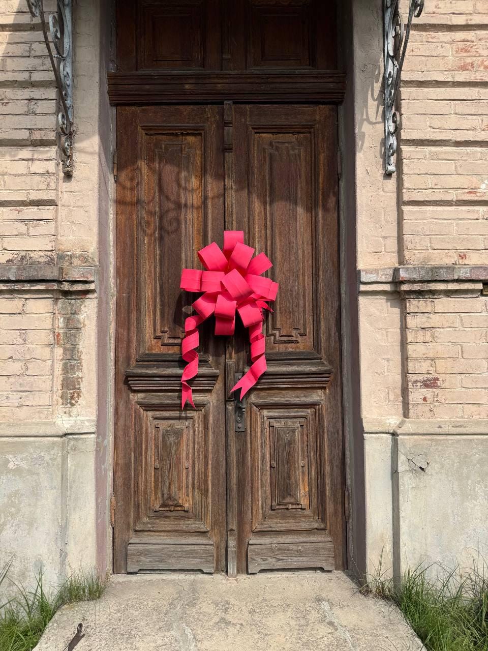 May include: A weathered, wooden double door with a bright pink bow. The door has detailed paneling and a dark finish. The bow is centered on the door and has long, flowing ribbons. The door is set in a brick building.