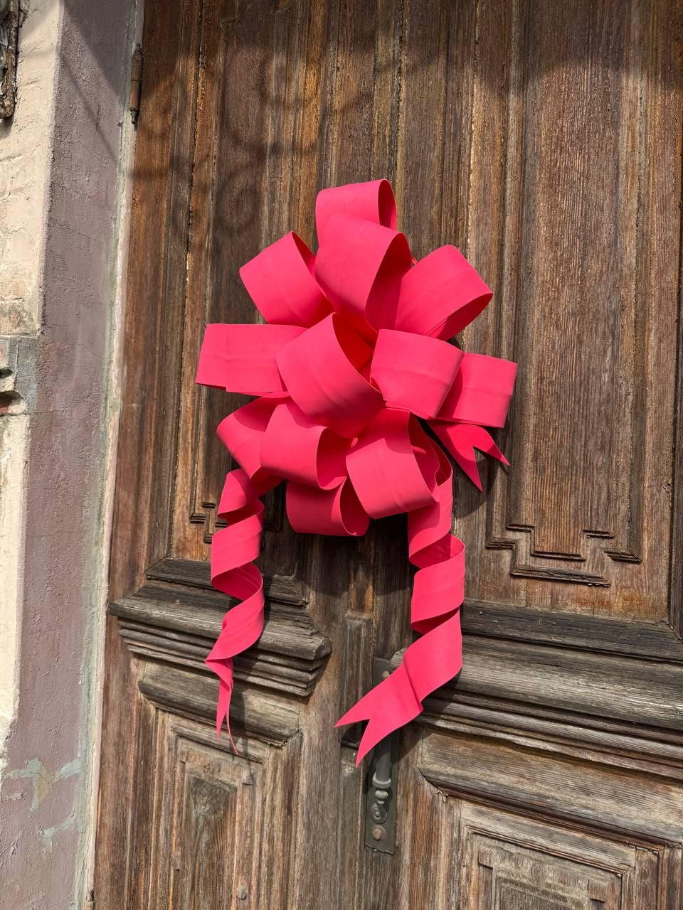 May include: A large, vibrant pink bow with long, curling streamers is affixed to a weathered wooden door. The bow's multiple loops and folds create a full, decorative appearance, perfect for festive occasions.