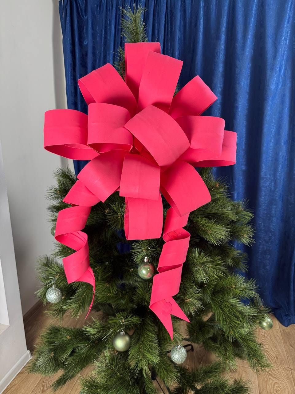May include: A vibrant pink bow adorns the top of a green Christmas tree. The bow features large, looped ribbons and cascading streamers. Silver and green ornaments are scattered throughout the tree.