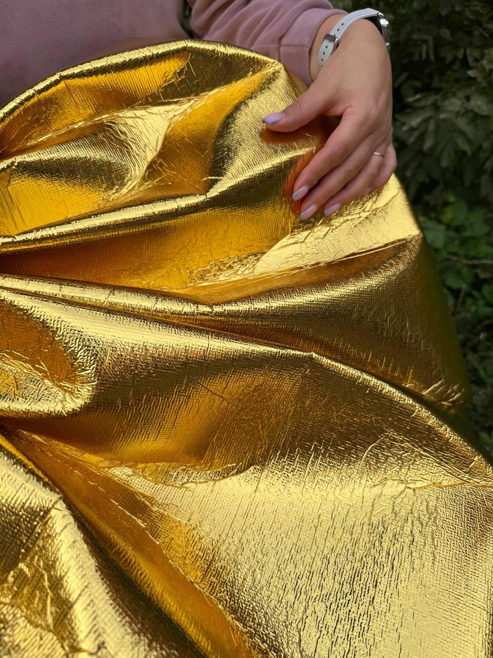 May include: A large, golden bow made of textured, metallic fabric. The bow is folded and gathered, creating a voluminous shape. The shiny material reflects light, highlighting the folds and curves of the bow. The bow is held by a person wearing a pink sweater.