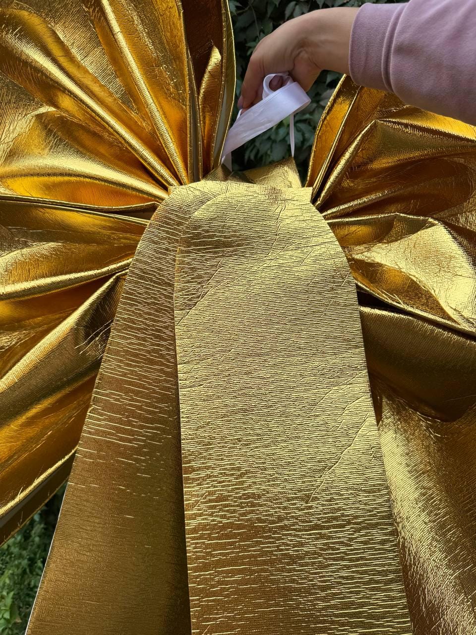 May include: A large, golden bow made of textured fabric. The bow has multiple layers and a shiny finish. A white ribbon is attached to the bow. The bow is likely used for decoration.