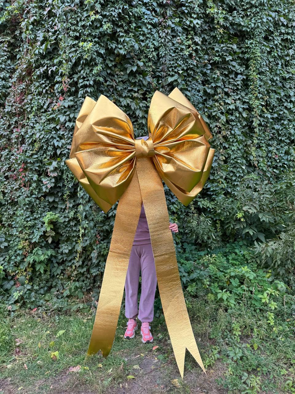 May include: A large, golden bow with long, flowing ribbons. The bow is made of a shiny, metallic material and is positioned in front of a green, leafy background. The bow's size suggests it is for a large gift or decoration.