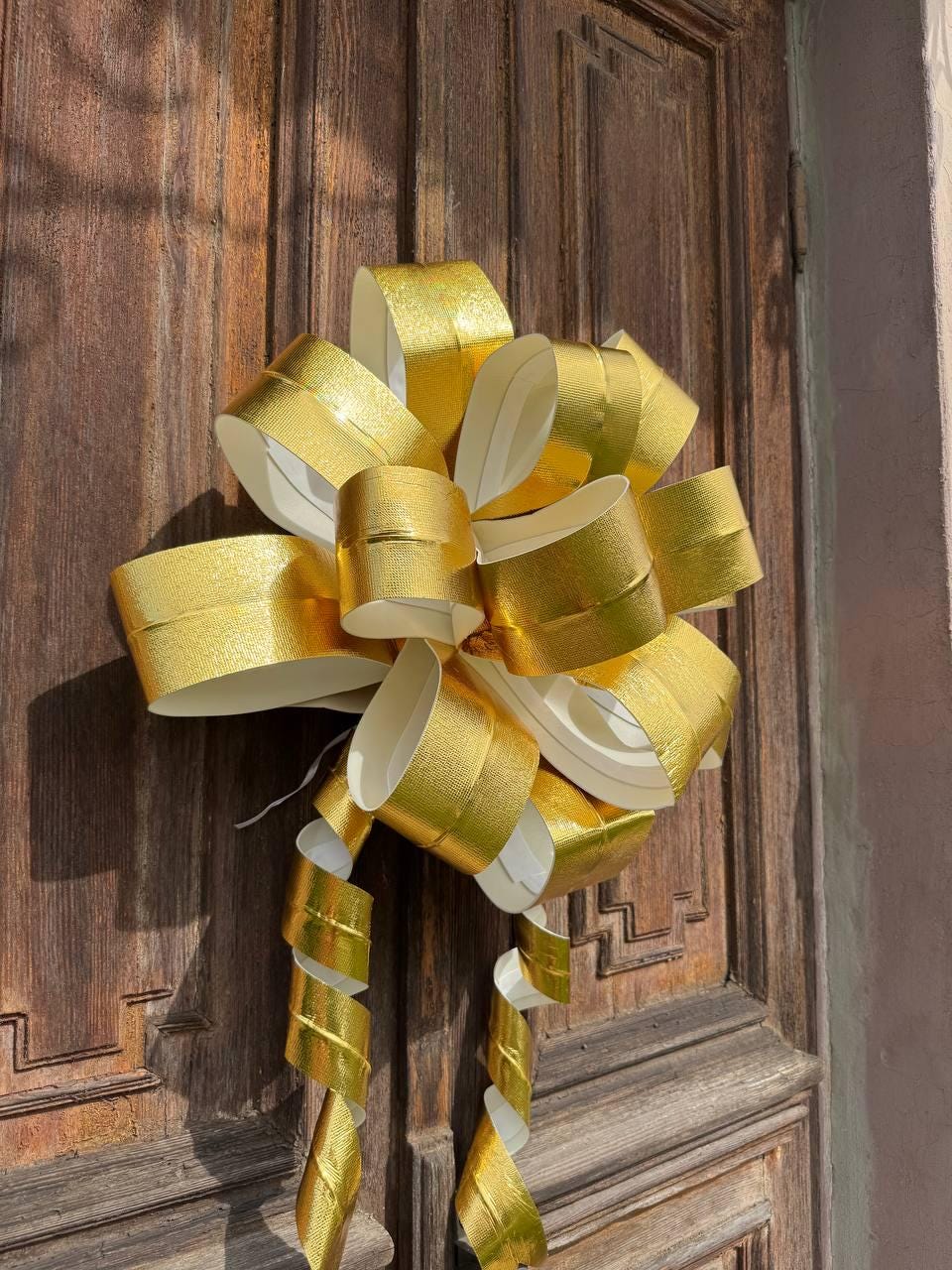 May include: A large, decorative gold bow with a metallic sheen. The bow is made of layered ribbons and has long, curled streamers. It is attached to a wooden door, suggesting it is for decoration or a gift.