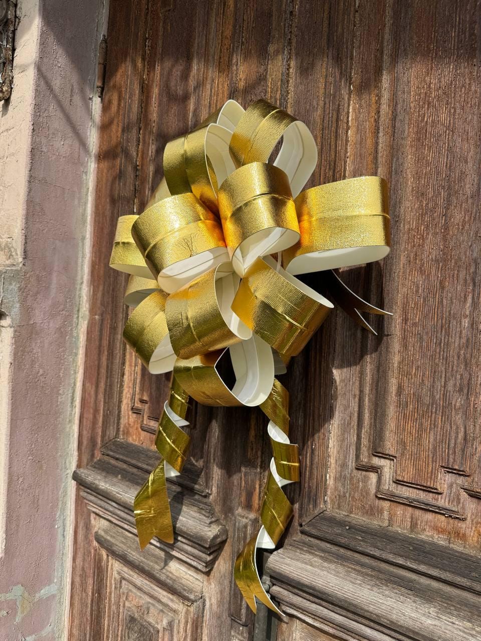 May include: A large, decorative gold bow with a metallic sheen, attached to a wooden door. The bow features multiple loops and cascading ribbons, creating a festive and elegant appearance. The bow's color is a bright, shiny gold.