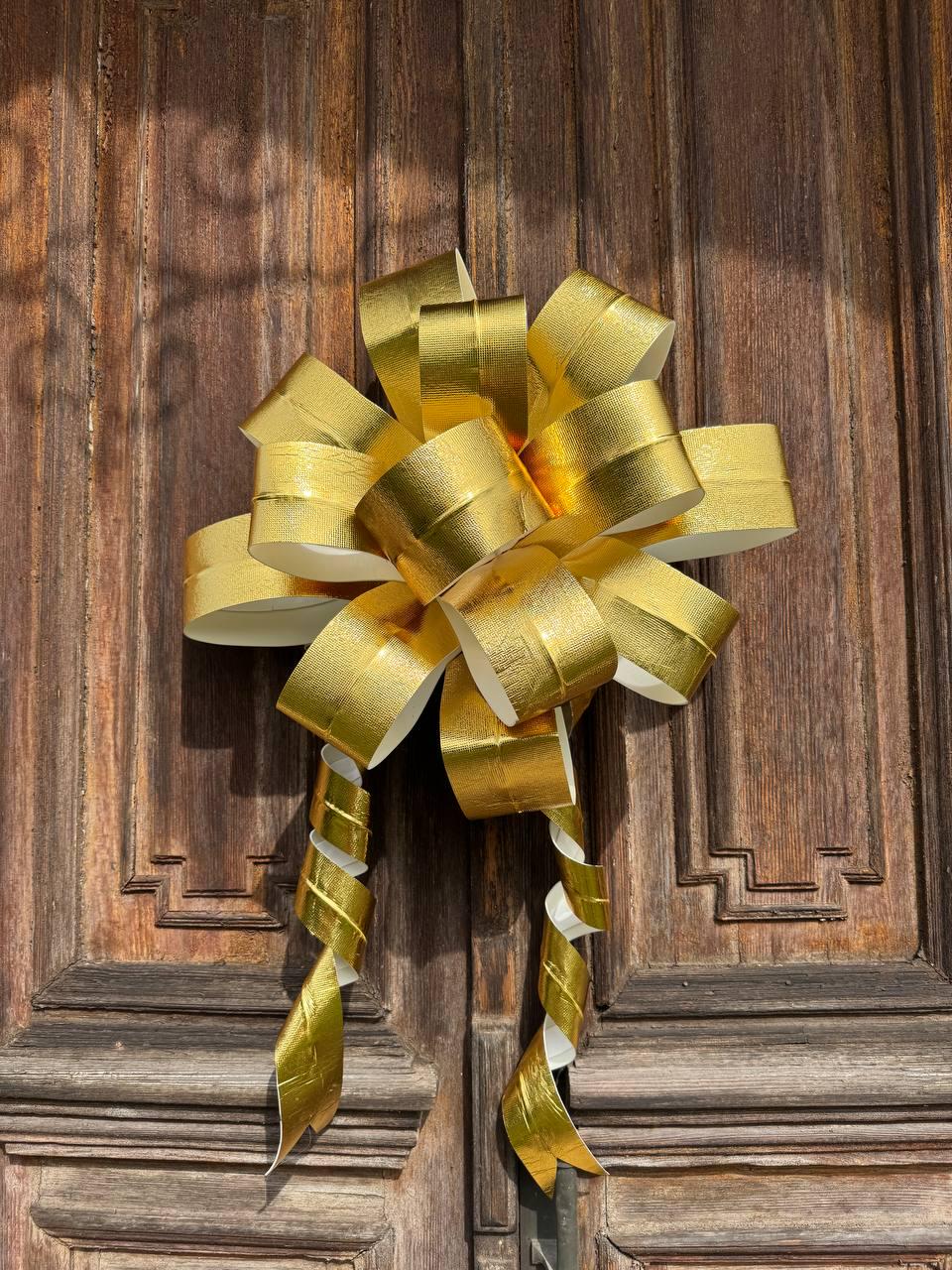 May include: A large, decorative gold bow with spiraling ribbon tails. The bow is made of shiny, metallic gold material and is set against a dark, wooden door. The bow is likely used for gift wrapping or decoration.
