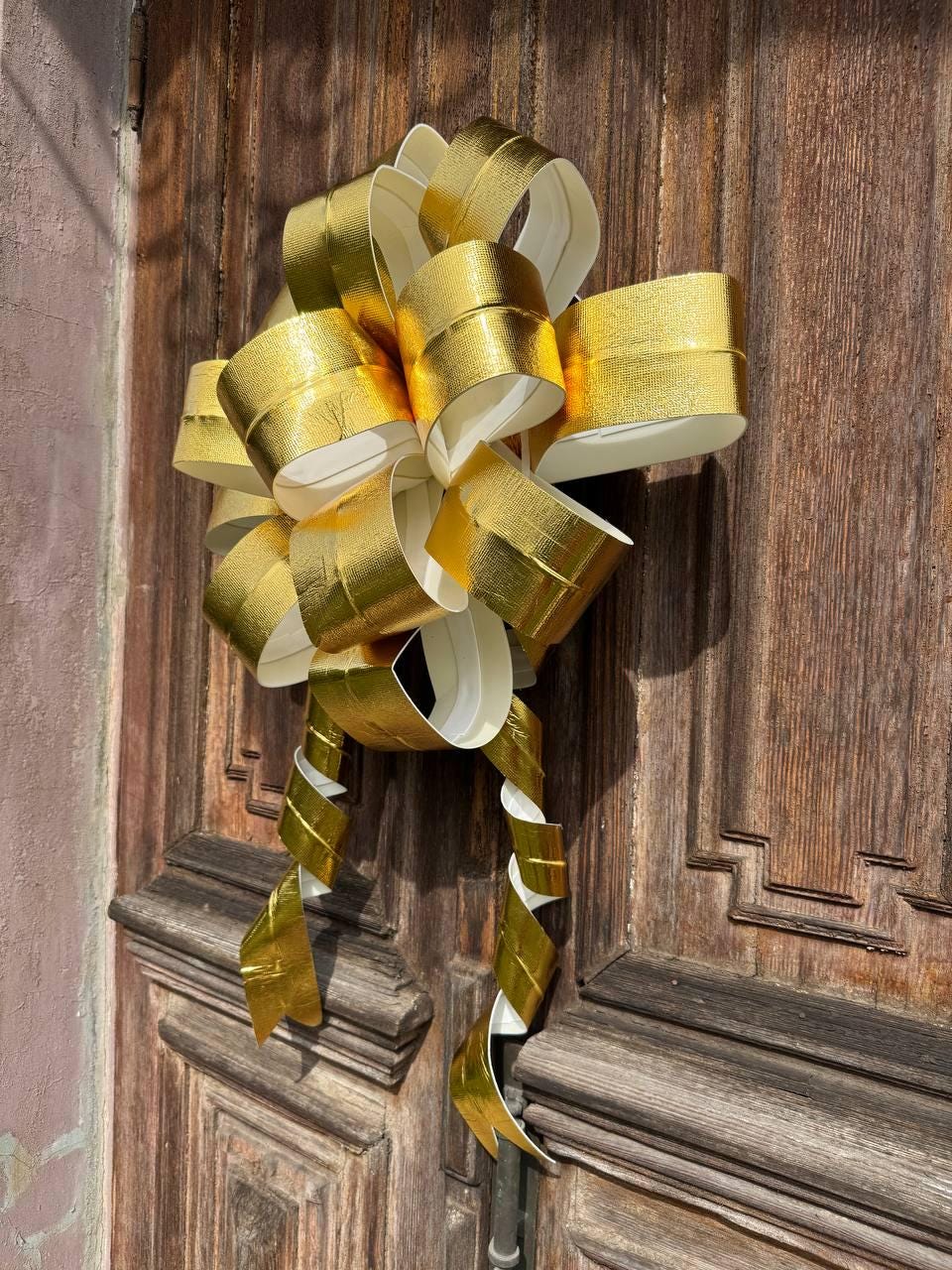 May include: A large, decorative gold bow with a metallic sheen, attached to a weathered wooden door. The bow features multiple loops and long, curling streamers, creating a festive and eye-catching display. The bow's color is a bright, shiny gold.