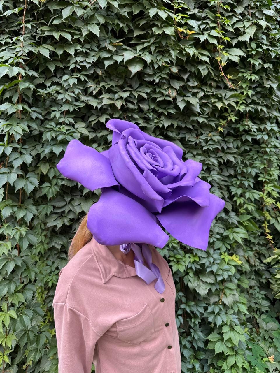 May include: A large, vibrant purple rose headpiece. The oversized floral design covers the head and face, with a soft, flowing ribbon. The person is wearing a light pink button-down shirt. The background is a wall of green leaves.