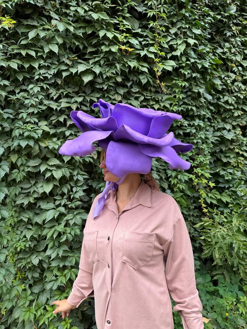 May include: A large, vibrant purple flower hat with oversized petals. The hat is worn by a person wearing a pink button-down shirt. The background is a lush green wall of leaves.