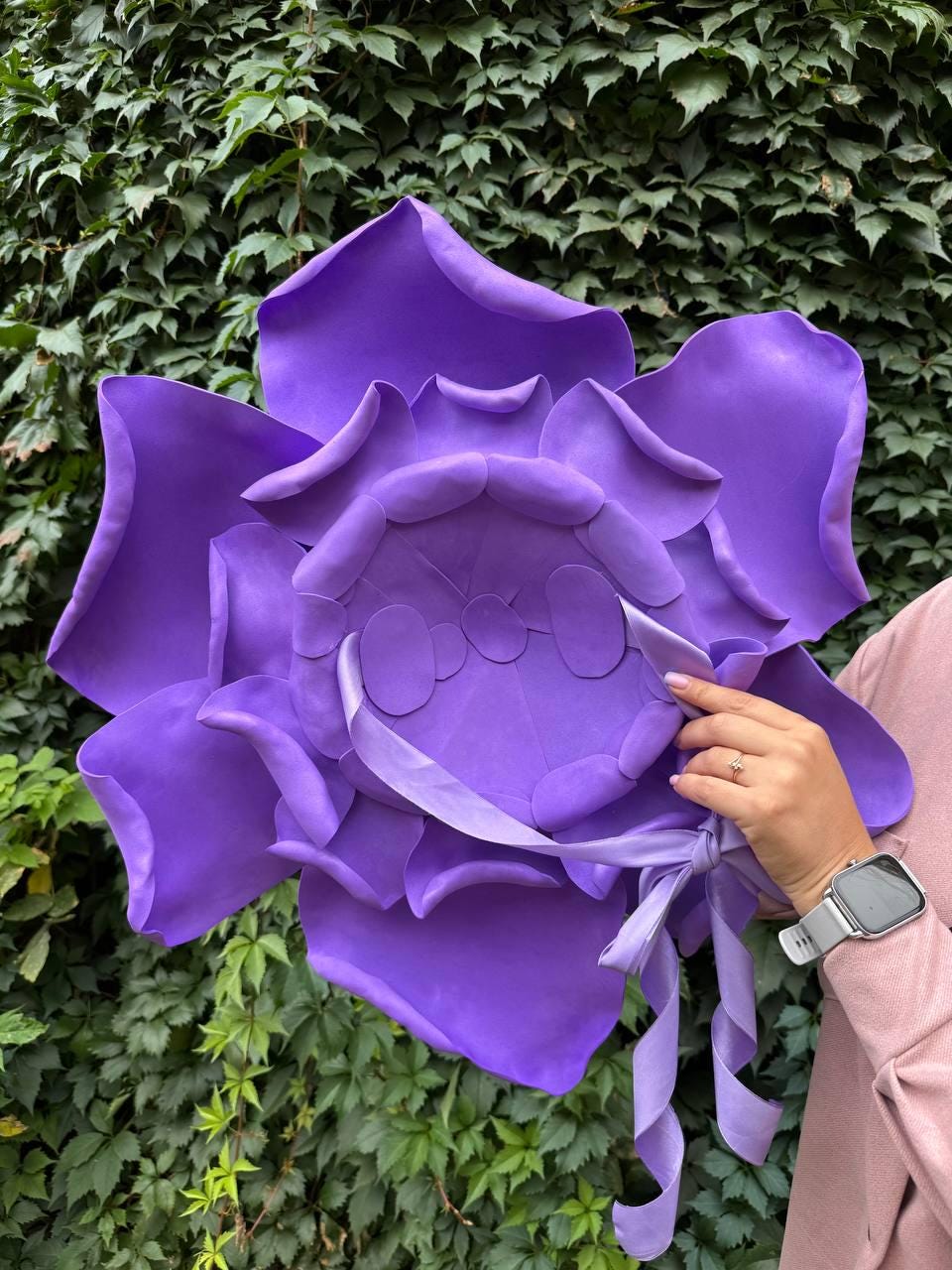 May include: A large, vibrant purple flower with layered petals and a ribbon tie. The flower is held against a backdrop of green foliage. The petals have a soft, velvety texture.