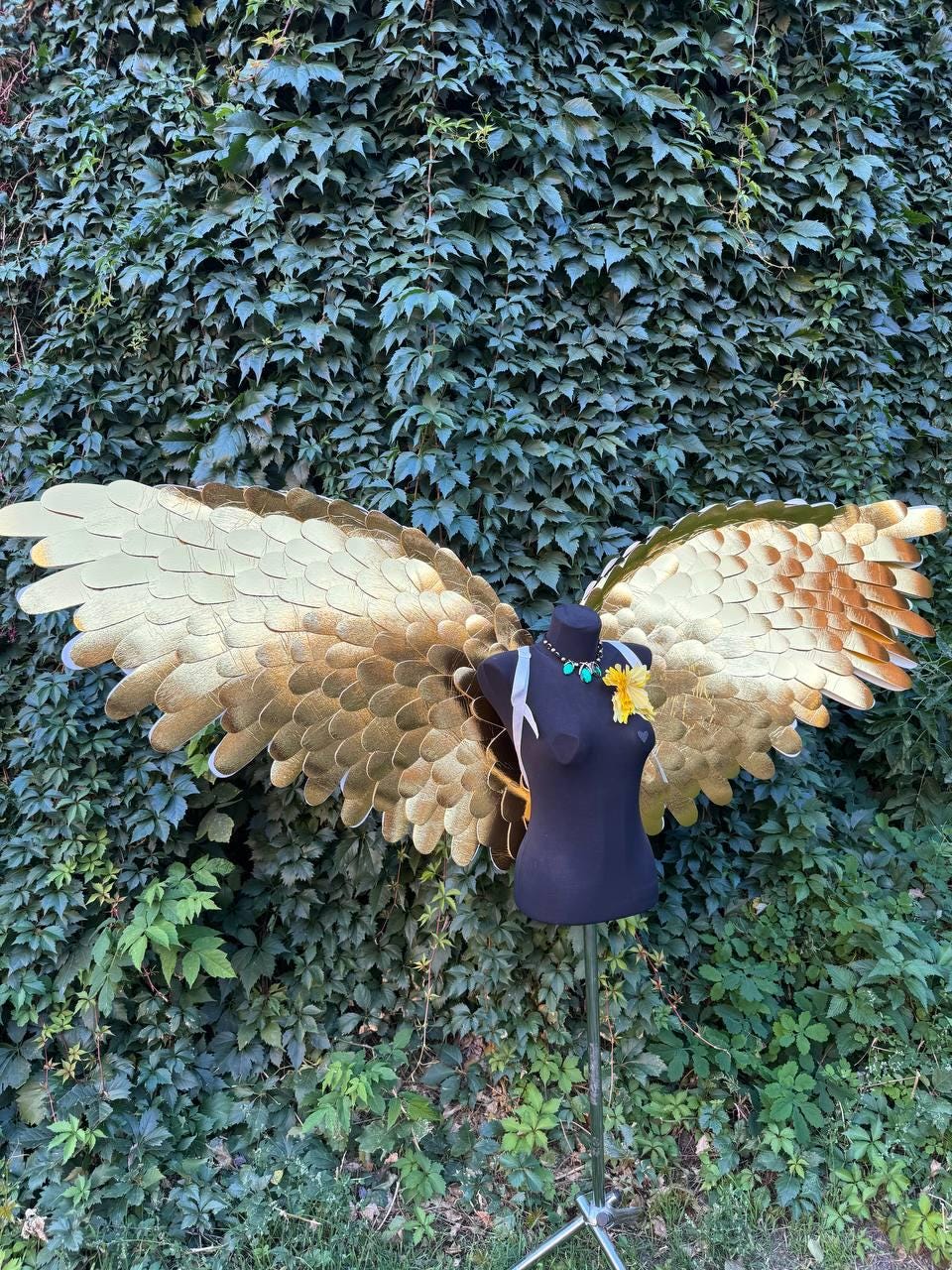 May include: Gold-colored costume wings with a layered feather design are displayed on a black mannequin. The wings are attached to the mannequin with white ribbons. A necklace and yellow flower adorn the mannequin's neck. The background is a wall of green leaves.