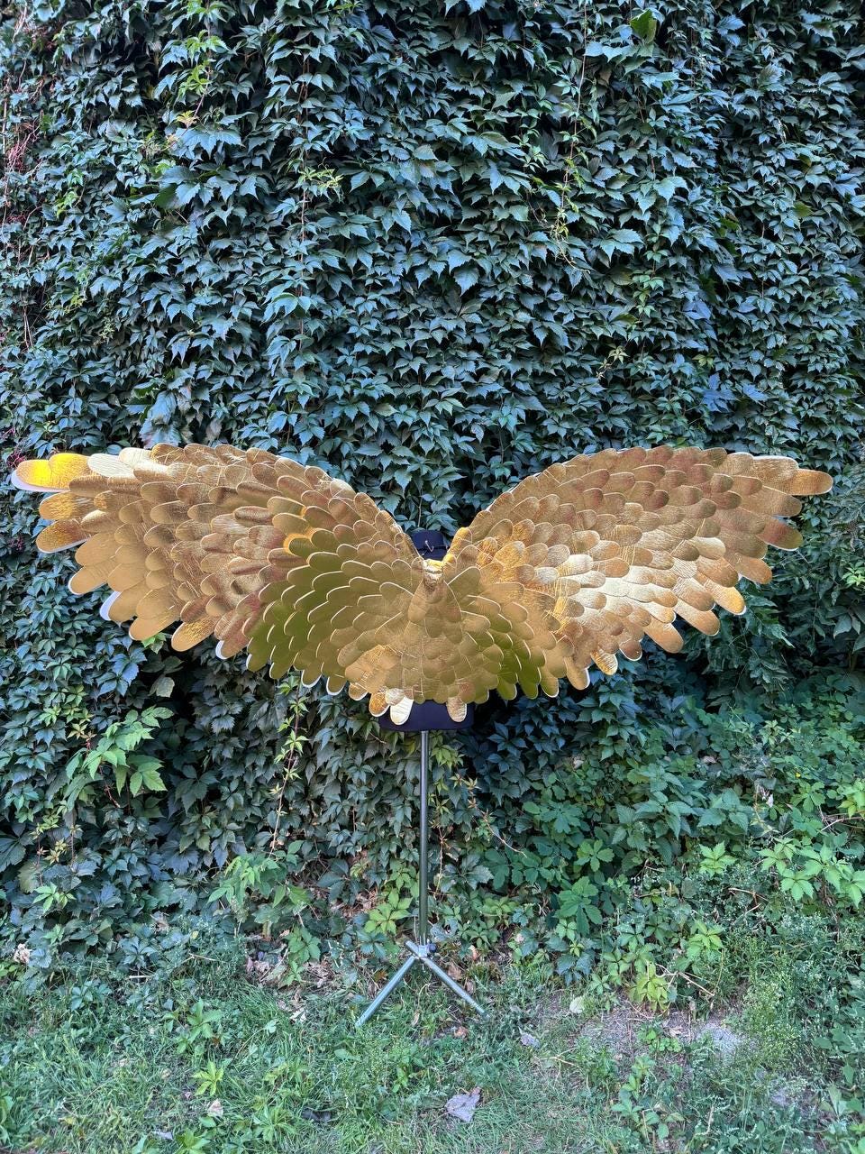 May include: Gold-colored, layered feather wings, displayed on a black stand against a backdrop of lush green foliage. The wings have a wide span, with a gradient of gold hues. The stand is supported by a tripod base.