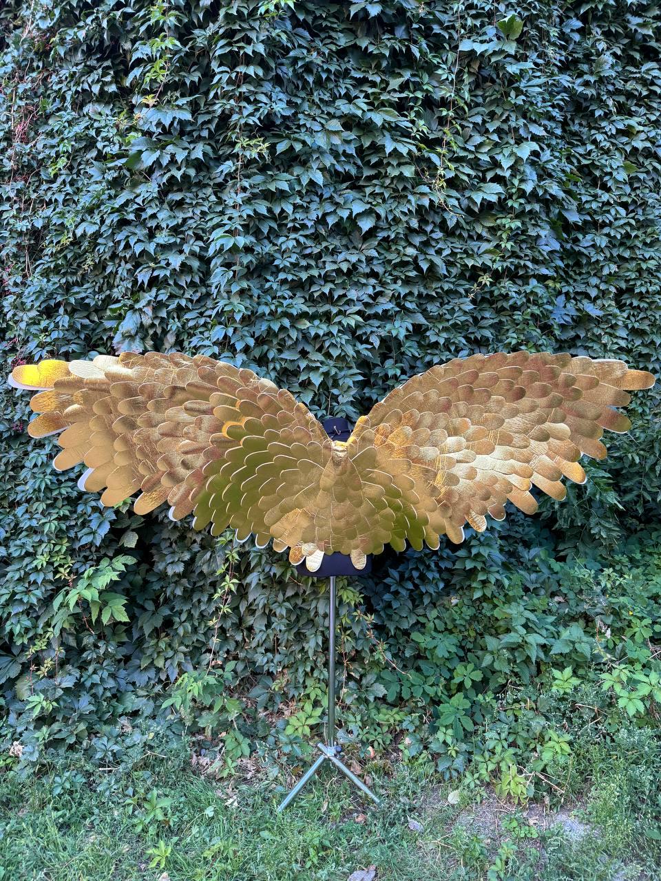 May include: A pair of large, golden-colored wings, possibly for a costume or decoration, are displayed against a backdrop of lush green foliage. The wings are made of overlapping, textured segments, creating a feathered appearance. They are mounted on a black stand.