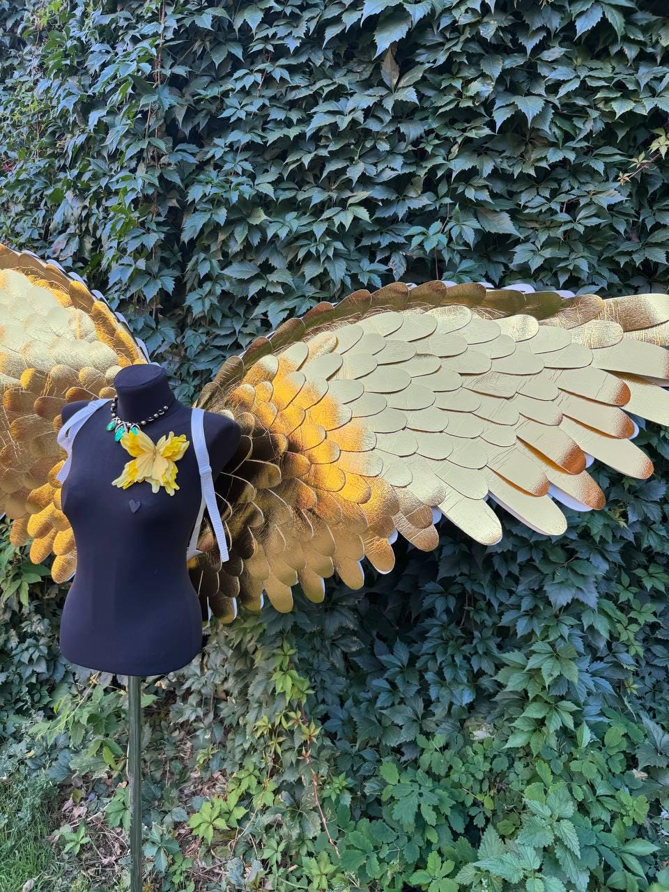 May include: Gold-colored costume wings with a layered feather design. The wings are displayed on a black mannequin with a yellow flower and necklace. White straps are attached to the wings. The background is a wall of green leaves.