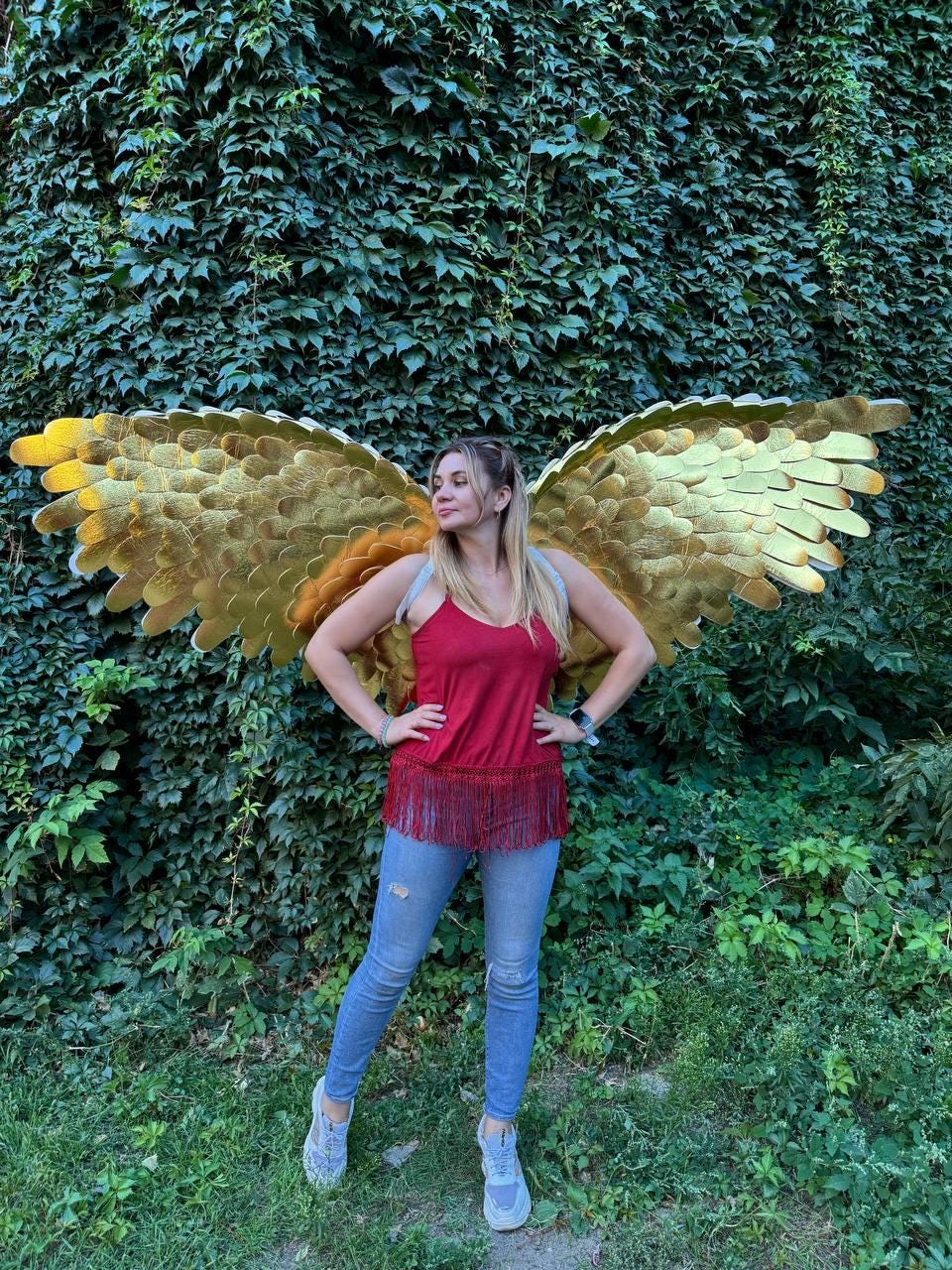 May include: A woman wearing a red top with a fringe hem and blue jeans stands in front of a wall of green foliage. Large, golden angel wings are attached to her back, with detailed feathering. The wings are the focal point of the image.