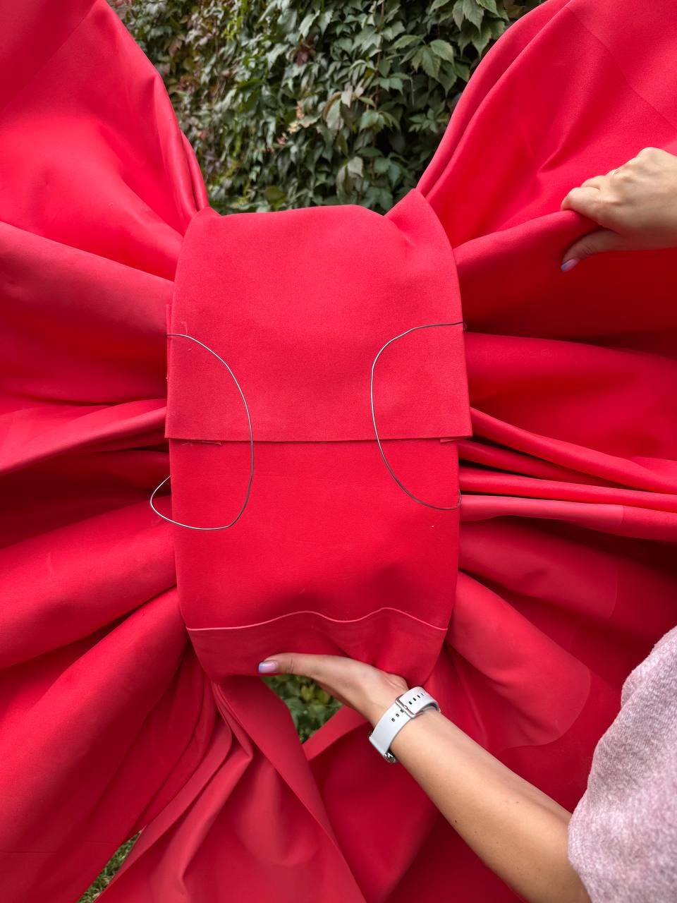 May include: A large, vibrant red bow made of fabric. The bow is being held by two hands, with a white watch visible on one wrist. The bow's design features a central rectangular section with wire detailing.