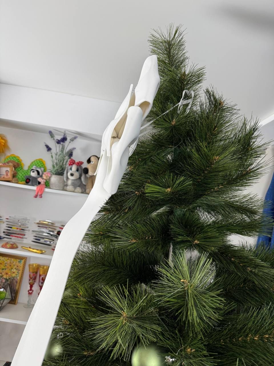 May include: A white Christmas tree ribbon is draped over a green artificial Christmas tree. The ribbon is made of a soft, flowing material. The tree is decorated with green ornaments. A shelf in the background displays various decorative items.