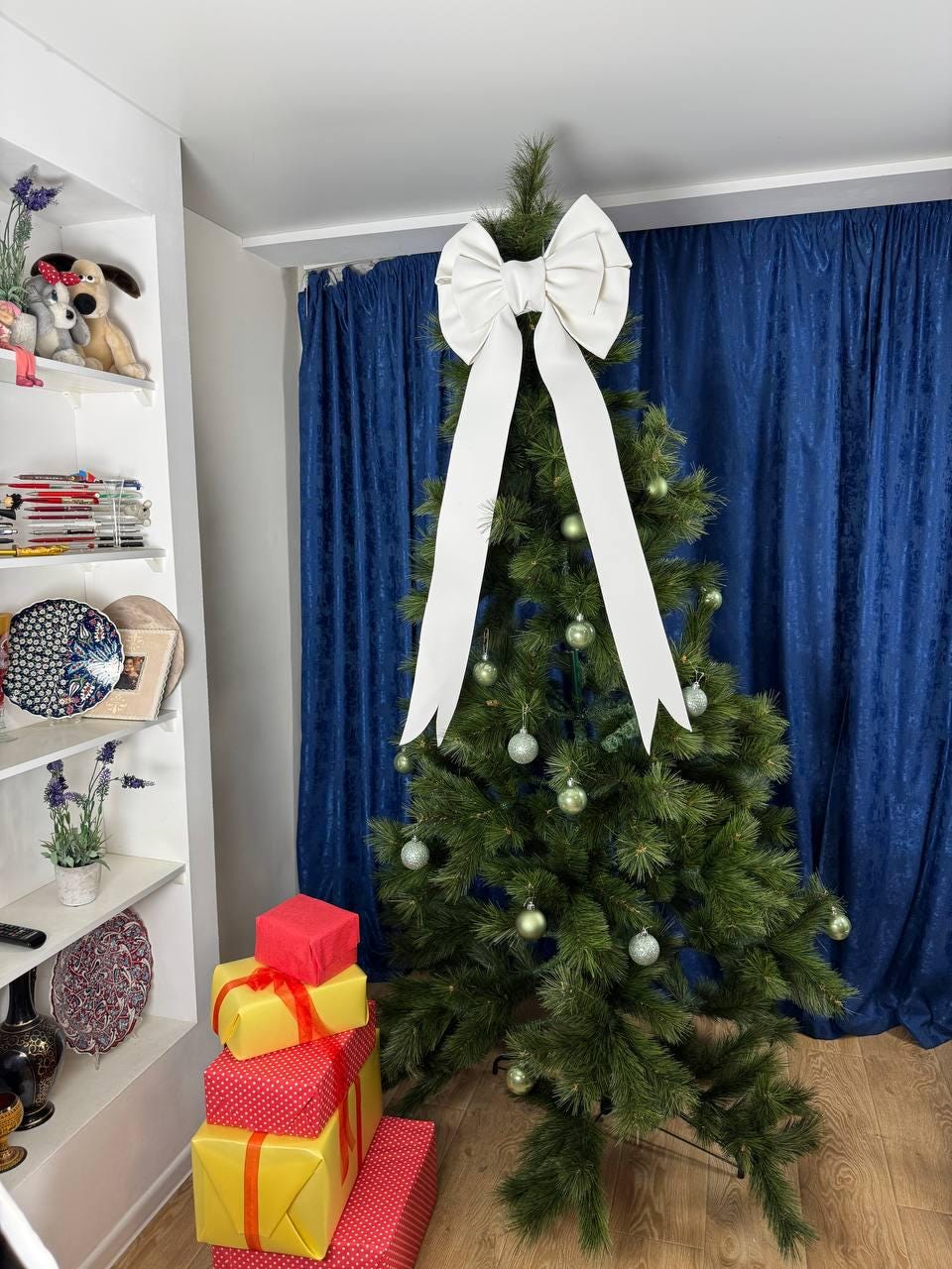 May include: A decorated Christmas tree with a large white bow at the top and silver ornaments. Wrapped gifts in red and yellow are at the base of the tree. The tree is set against a blue curtain backdrop.