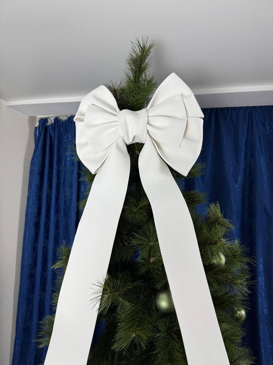 May include: A large, white bow adorns the top of a Christmas tree. The bow is made of a smooth material and has long, flowing tails. The tree is decorated with green ornaments and set against a blue curtain backdrop.