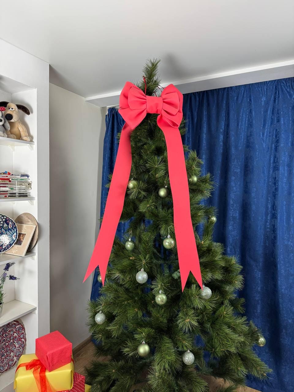 May include: A decorated Christmas tree with a large red bow at the top and long, flowing ribbons. The tree is adorned with silver and green ornaments. The background includes a blue curtain and a white shelf with decorative items.
