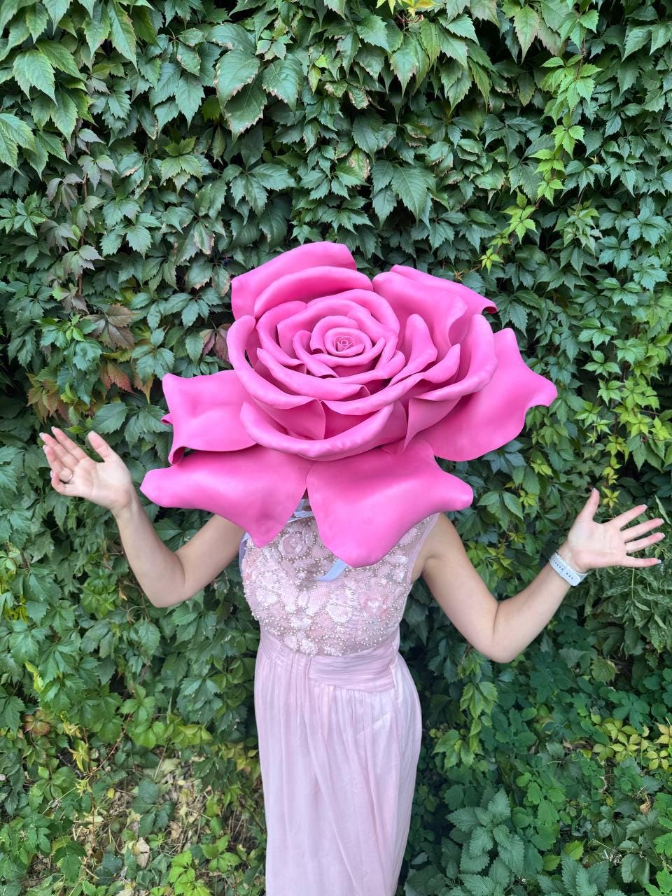 May include: A person wearing a large, vibrant pink rose headpiece and a light pink dress. The rose is the focal point, with detailed petals. The dress has a beaded bodice and a flowing skirt. The background is a lush green wall of leaves.