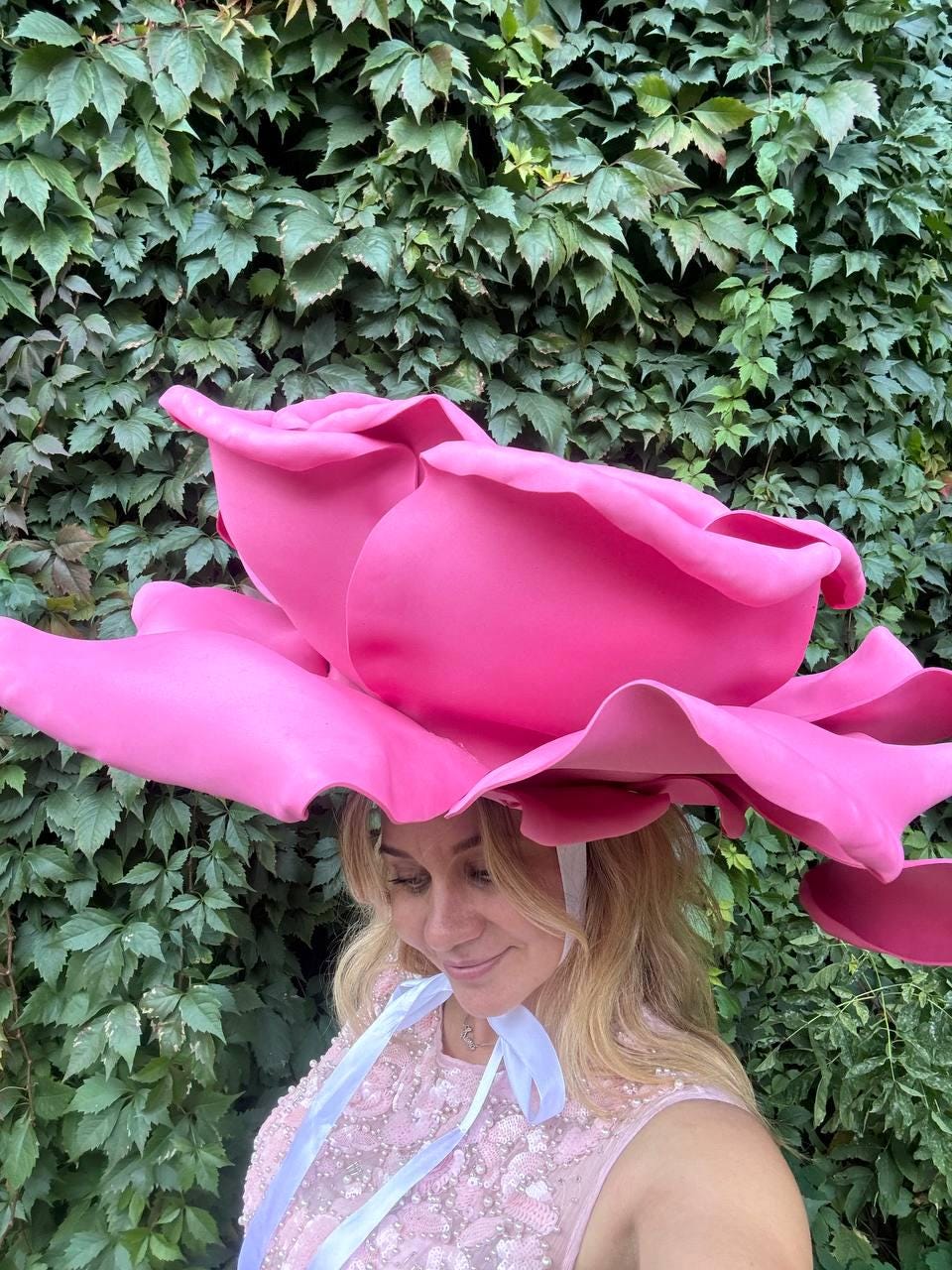 May include: A large, vibrant pink rose-shaped hat with a wide brim. The hat is made of a soft material and is secured with white ribbons. The background features lush green foliage, creating a striking contrast with the hat's color.