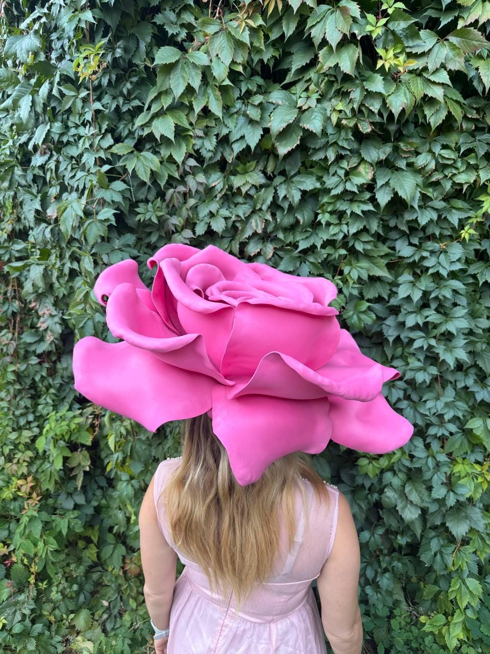 May include: A large, vibrant pink rose headpiece. The oversized flower is the focal point, with multiple petals. The person is wearing a pink dress. The background is a wall of green leaves.
