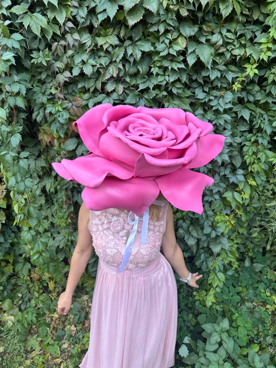 May include: A large, vibrant pink rose headpiece worn by a person in a light pink dress. The rose is the focal point, with detailed petals. The dress has a beaded bodice and a flowing skirt. A light blue ribbon is tied to the headpiece.