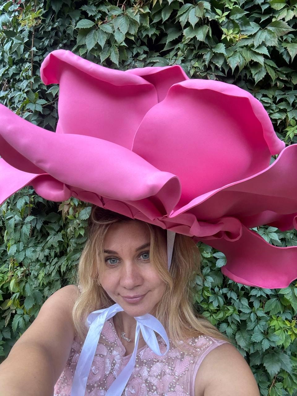 May include: A large, vibrant pink rose-shaped hat. The hat is worn with a white ribbon tied under the chin. The background features lush green foliage. The hat is a whimsical accessory.