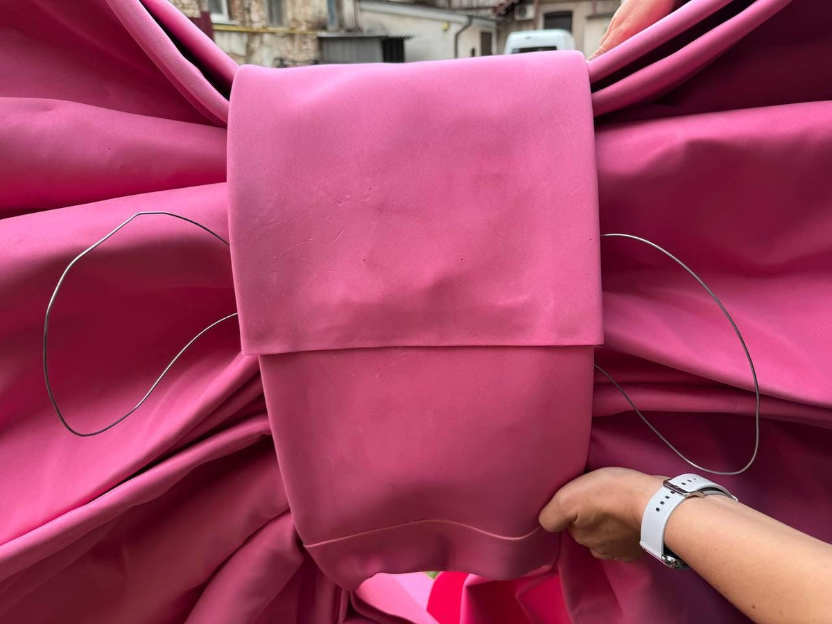 May include: A large, bright pink bow made of fabric. The bow has a rectangular center and two wire-like structures extending from the sides. The bow is held by a person wearing a white watch.