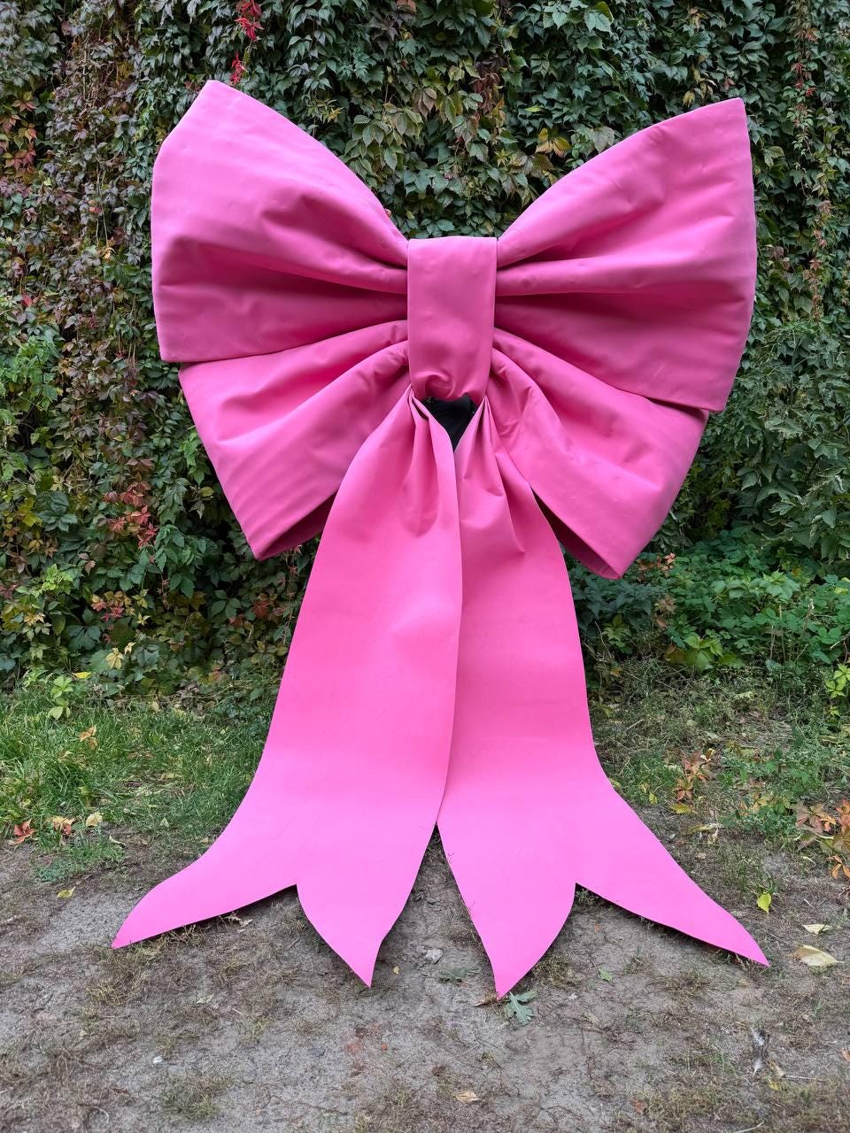 May include: A large, vibrant pink bow with long, flowing tails. The bow is made of a smooth, possibly satin-like material and is set against a backdrop of green foliage and grass. The bow's size suggests it's a decorative element.