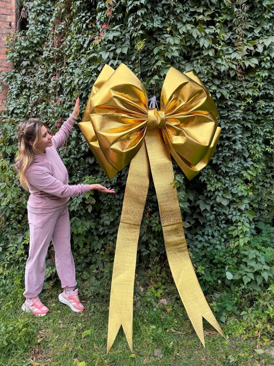May include: A large, golden bow with long, flowing ribbons is displayed against a backdrop of lush green foliage. The bow is made of a shiny, metallic material and appears to be a decorative element, possibly for a special event or holiday.