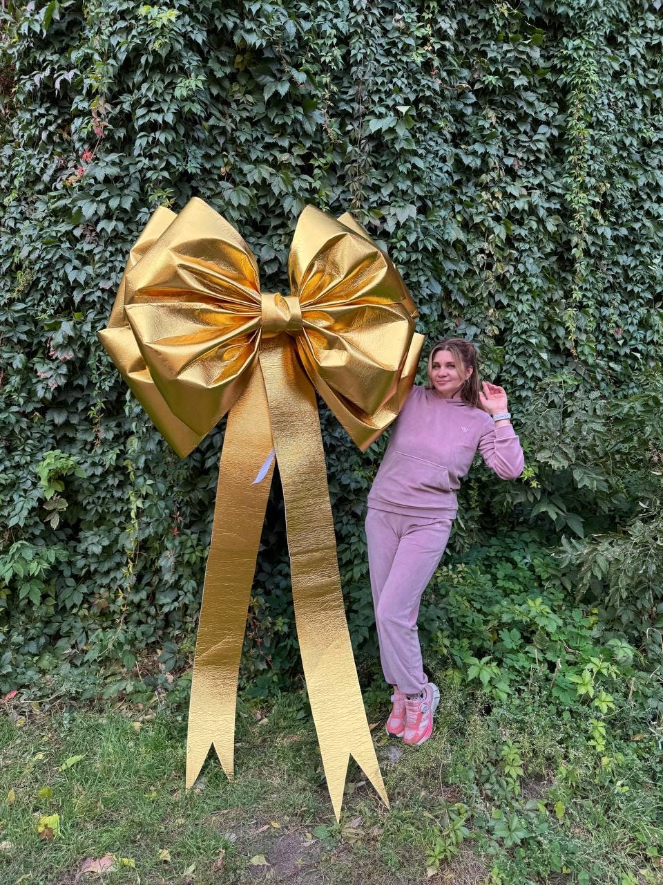 May include: A large, golden bow with long streamers. The bow is made of a shiny, metallic material and is positioned in front of a wall of green foliage. The bow is a festive decoration, suitable for celebrations and events.