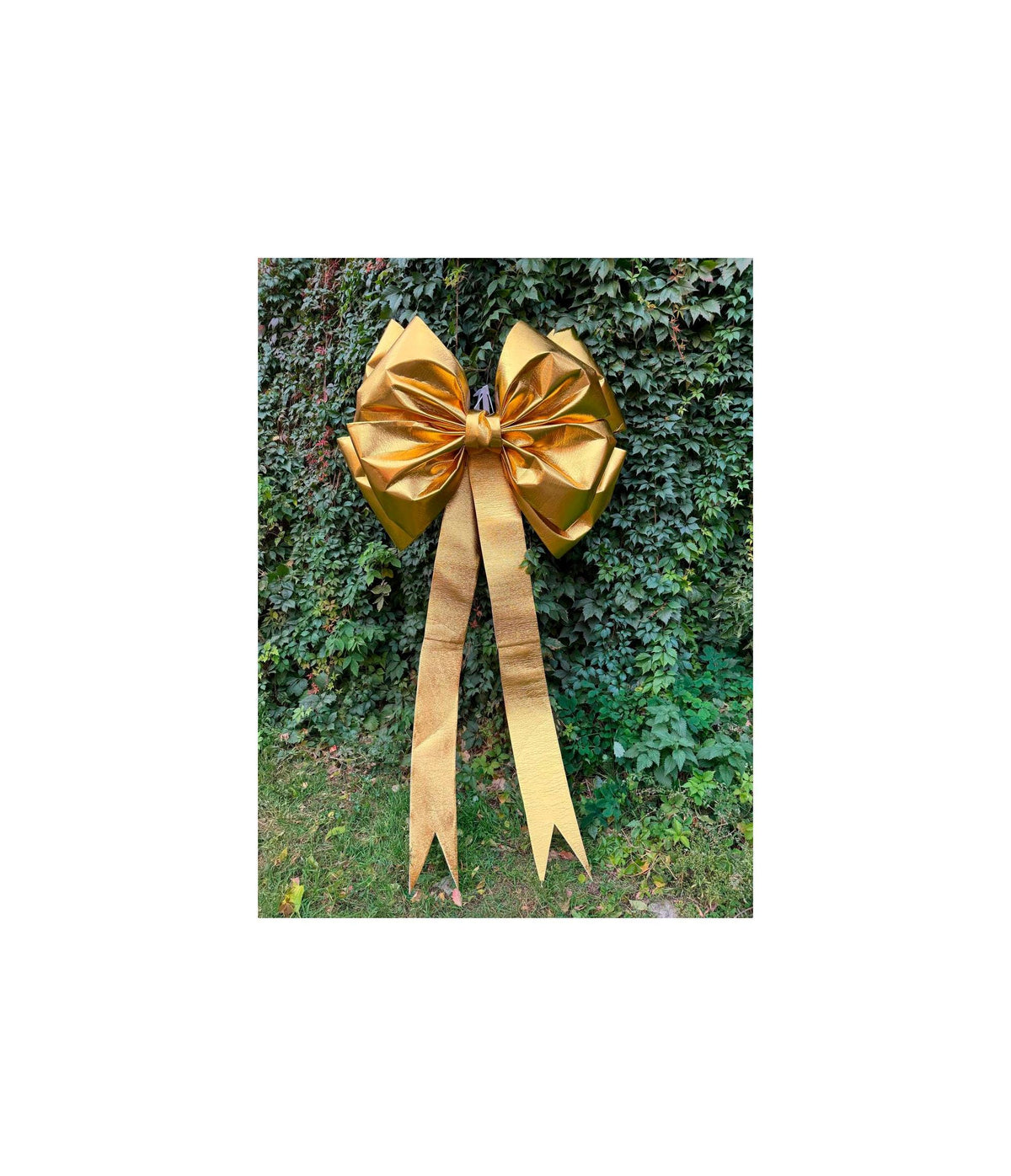 May include: A large, shiny gold bow with long, flowing ribbons. The bow is made of a metallic material and is displayed against a backdrop of green foliage. This decorative bow is suitable for festive occasions.