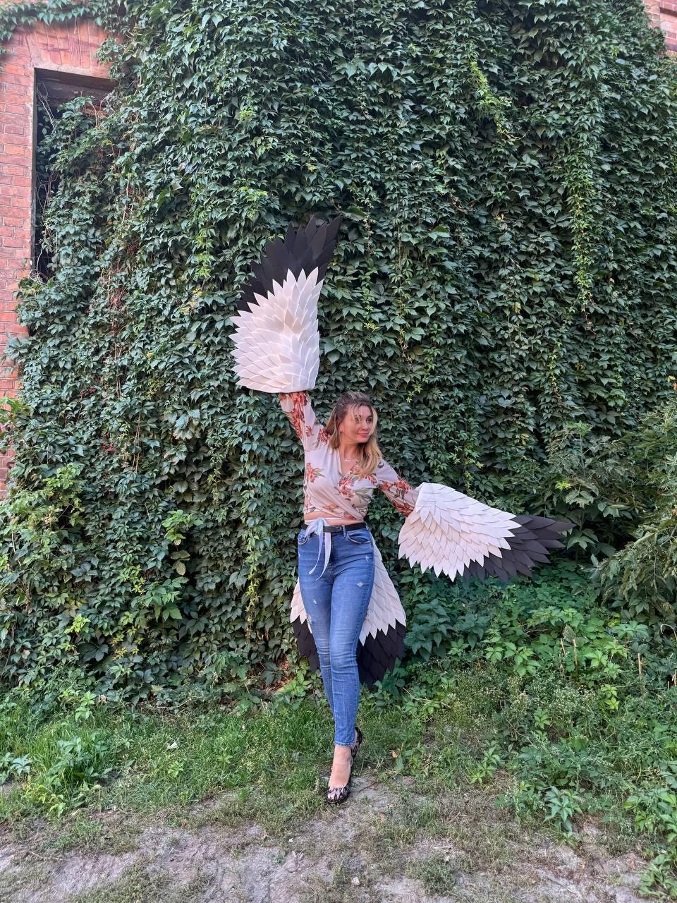 May include: A person wearing a floral top and blue jeans, posing with large, handmade white and black bird wings. The wings are spread wide against a backdrop of lush green foliage and a brick building.