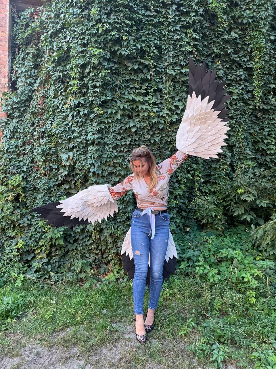 May include: A person wearing a costume with large, stylized bird wings. The wings are primarily white, black, and beige, with a layered, feathered appearance. The person is wearing a floral top and blue jeans.