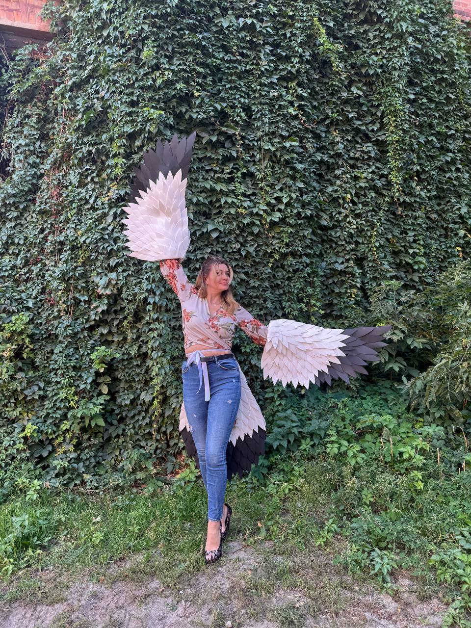 May include: A person wearing a floral top, blue jeans, and black heels, posing with large, white, black, and tan bird wings. The wings are made of layered, textured material and are spread wide against a backdrop of green foliage.