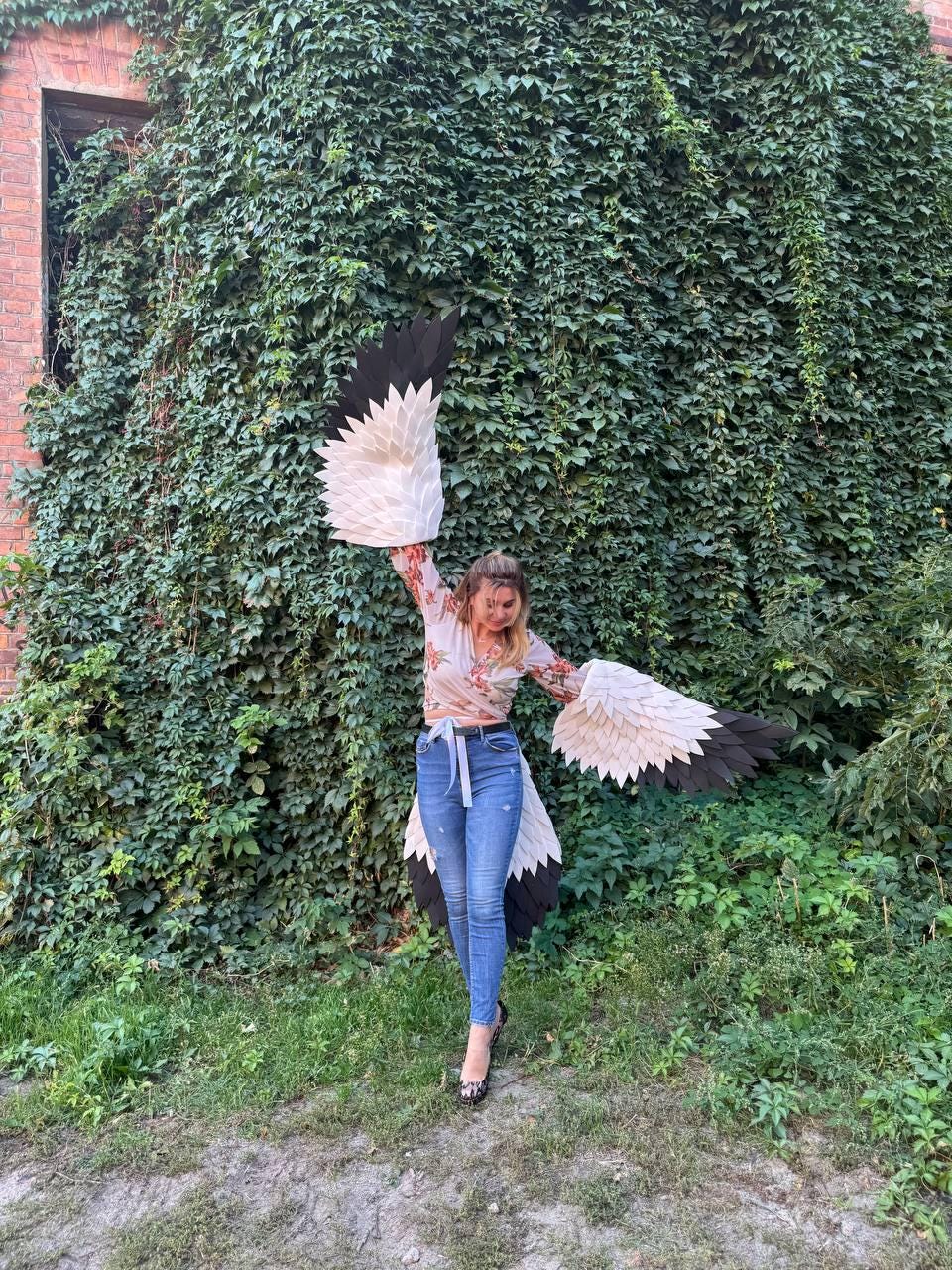 May include: A person wearing large, handmade bird wings with black and white feathers. The wings are spread wide against a backdrop of lush green foliage and a brick building. The person is wearing a floral top and blue jeans.