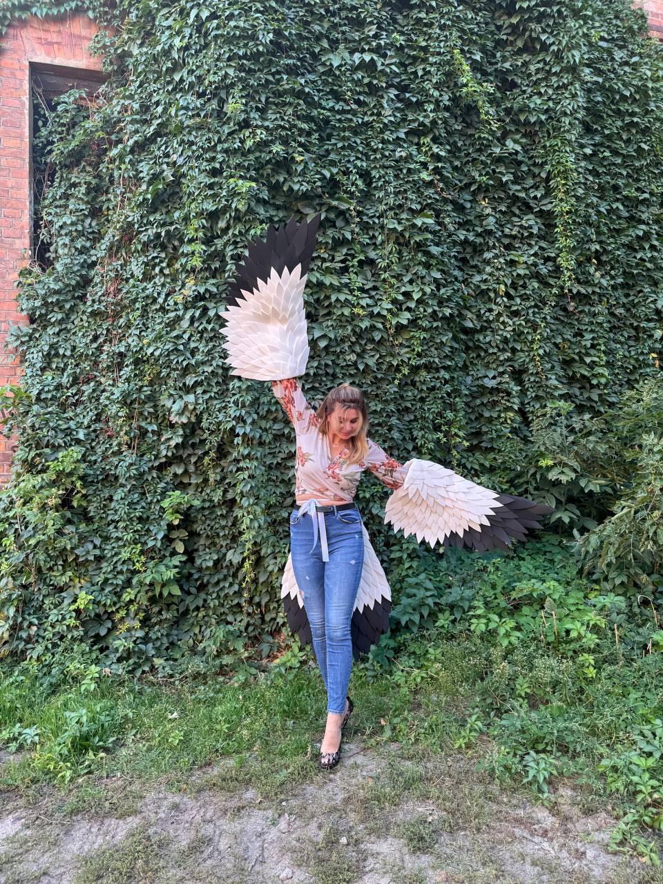May include: A person wearing a floral top and blue jeans with large, white, black, and beige bird wings. The wings are spread wide against a backdrop of lush green foliage and a brick building.