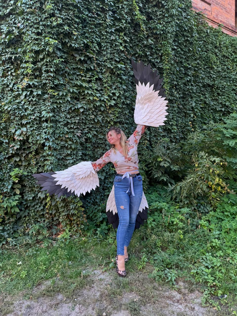 May include: A person wearing a floral top, blue jeans, and patterned shoes, posing with large, layered paper wings. The wings feature black, white, and cream-colored feathers, creating a striking visual effect against a backdrop of green foliage.