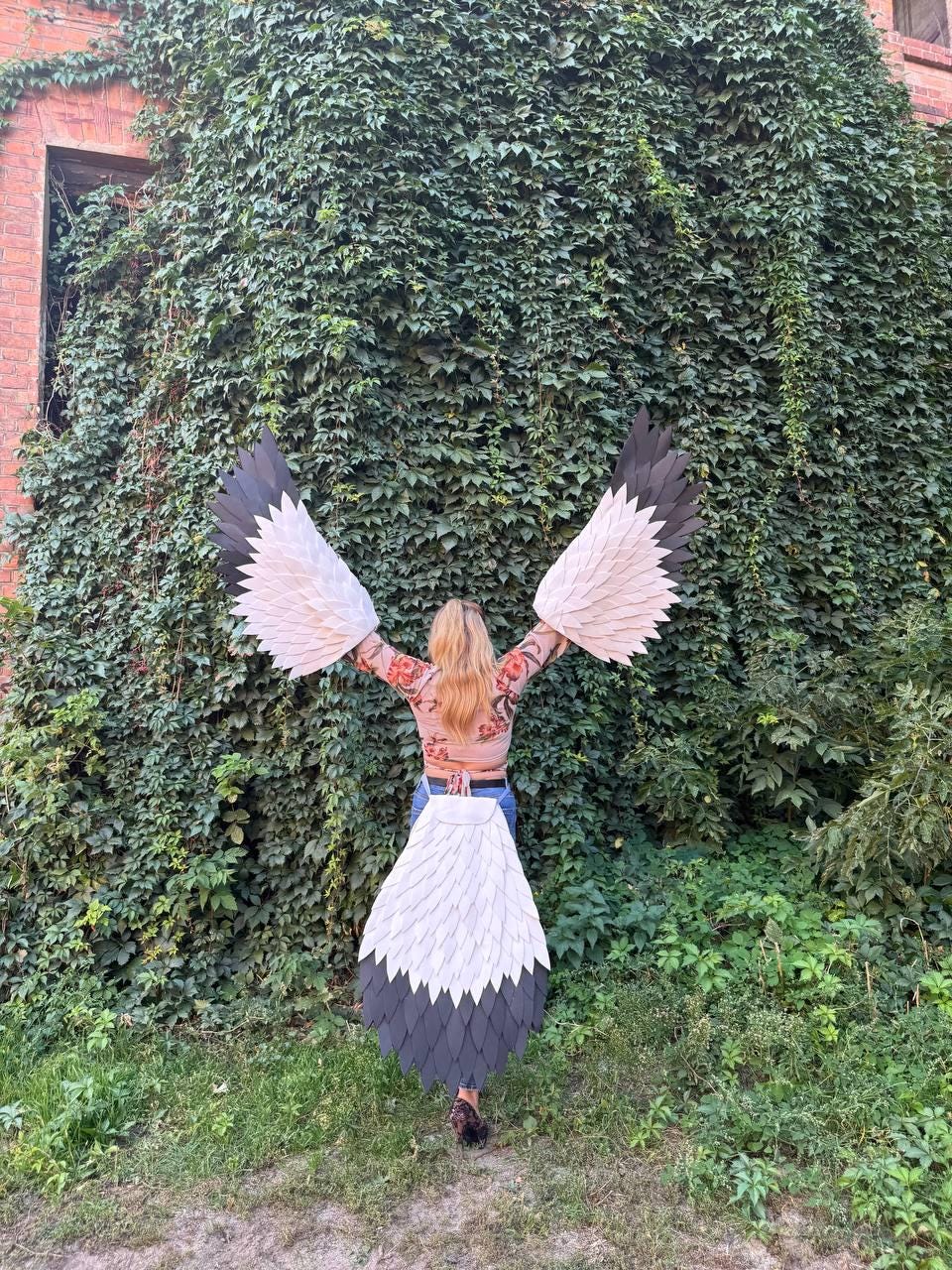 May include: A person wearing a costume with large, stylized wings. The wings are primarily white with black and gray accents, resembling a bird's plumage. The costume is worn outdoors against a backdrop of lush green foliage.