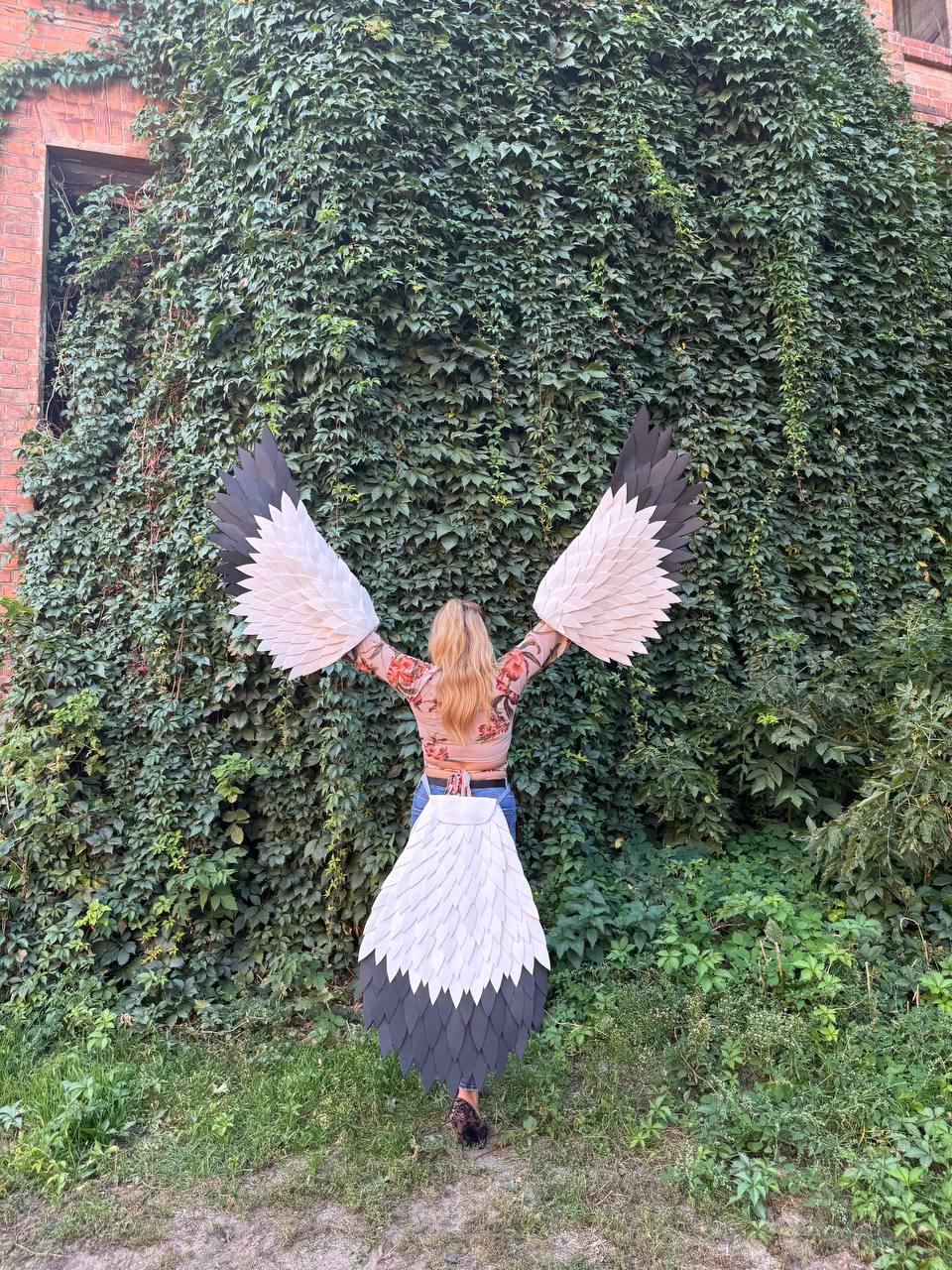 May include: A person wearing a costume with large, stylized wings. The wings are primarily white with black and gray accents, resembling a bird's plumage. The costume is worn outdoors against a backdrop of lush green foliage.
