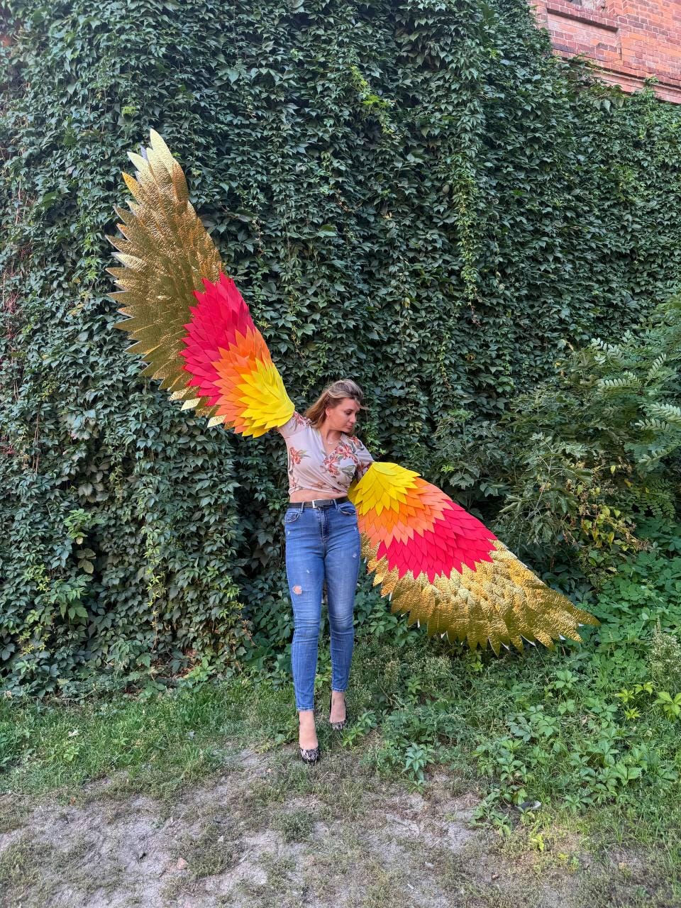 May include: A person wearing large, colorful wings with gold, red, orange, and yellow feathers. The wings are spread wide against a backdrop of green foliage and grass. The wings appear to be costume or decorative.